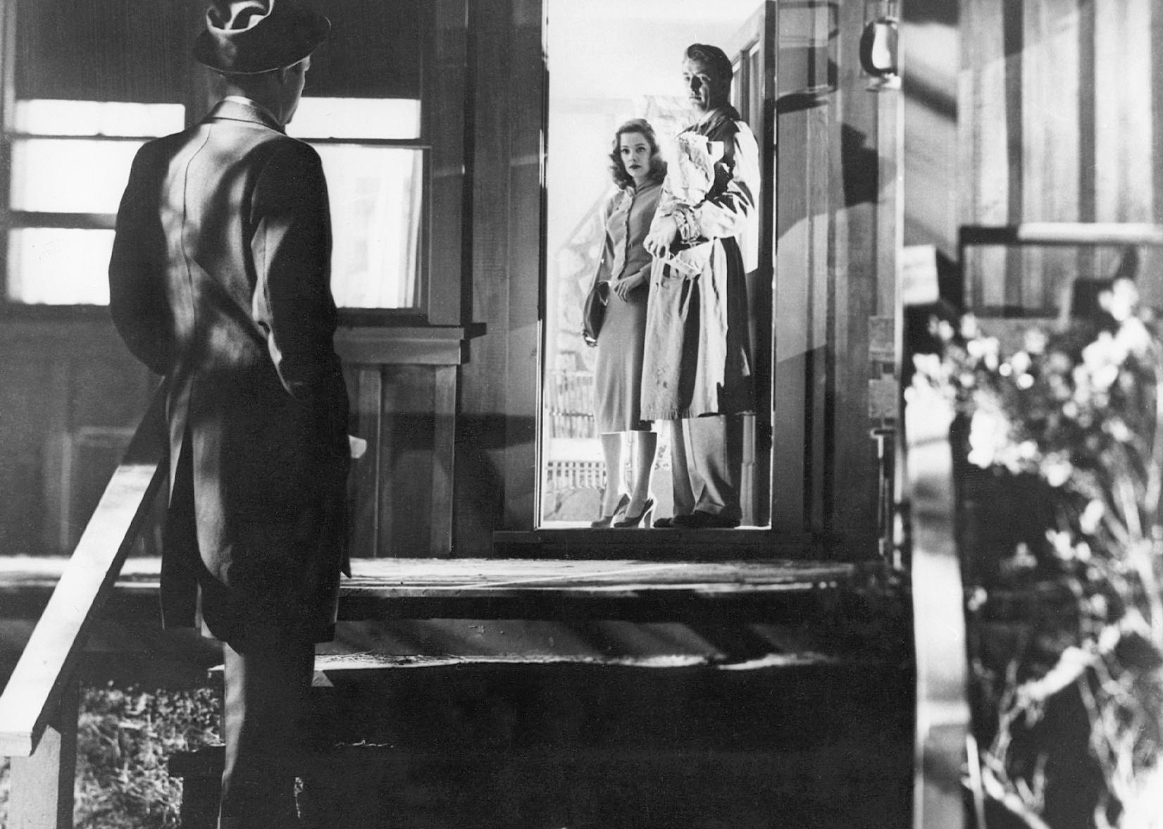 A nicely dressed man and woman stand in a doorway on a porch with a darkly lit figure at the bottom of the stairs looking at them.
