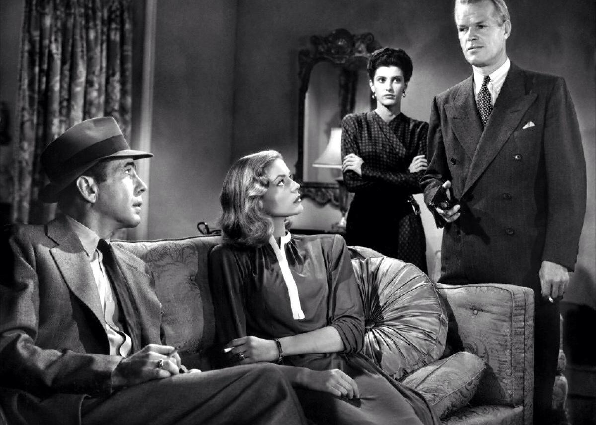A tall man in a suit points a gun at Lauren Bacall and Humphrey Bogart sitting on a couch with a woman looking on in the background.