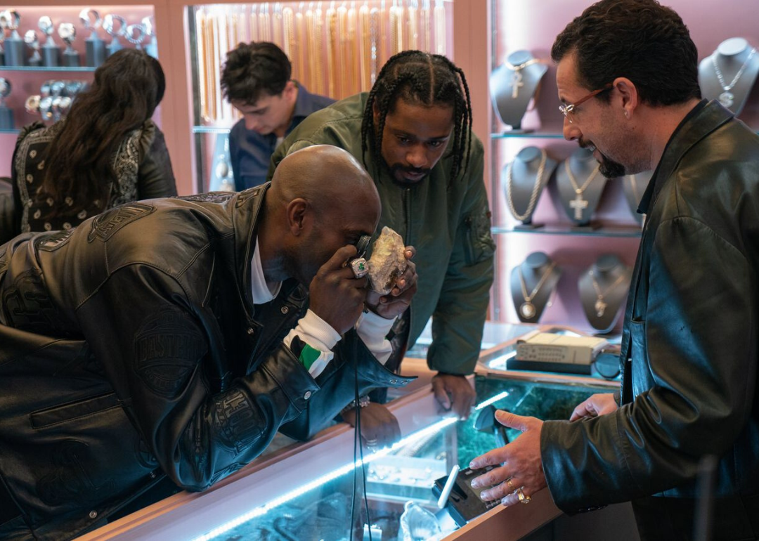 Adam Sandler stands behind a jewelry counter while two men look at jewels with a magnifying glass.