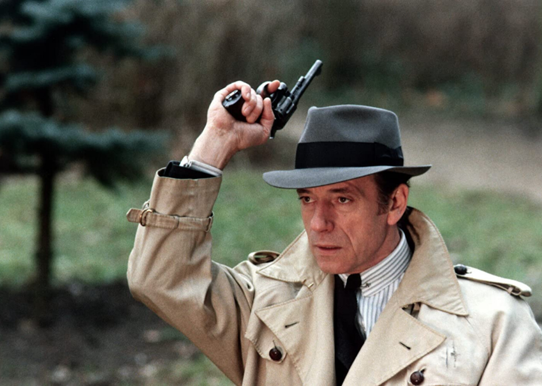 A man in a trench coat and hat with a gun in the air.