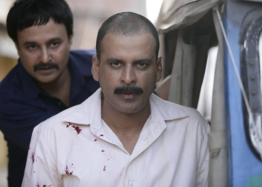 A man with a shaved head and blood spatter on his shirt stands next to a small car with another man looking on behind him.