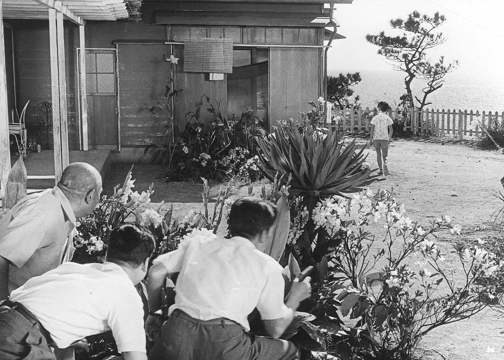 Three men hide in the bushes watching a house as a young boy goes around the side.