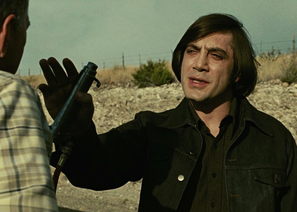 Javier Bardem holds a police baton towards a man's face.
