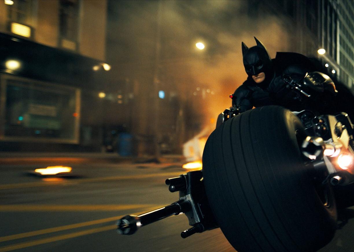 Batman rides a motorcycle with wide wheels racing through the city.