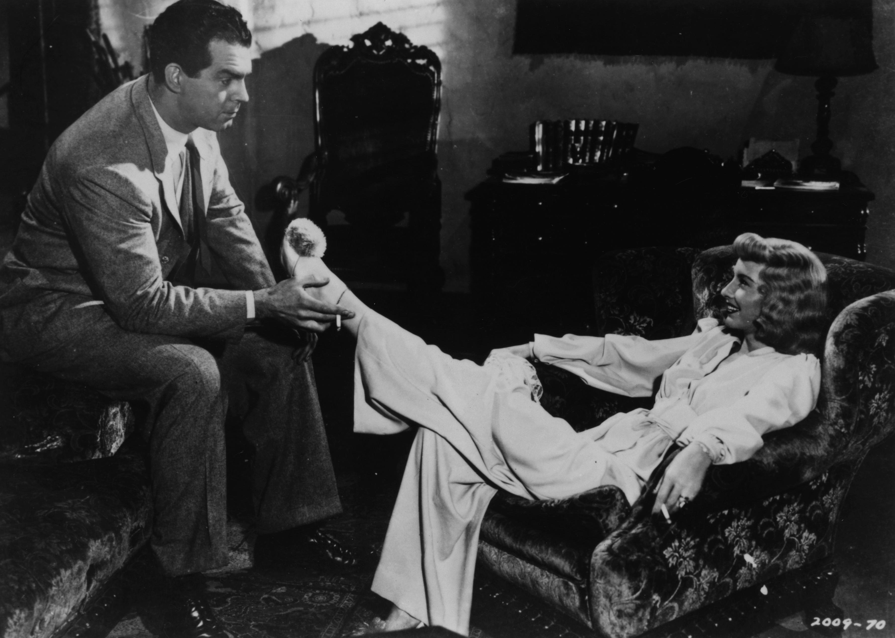 A man smoking a cigarette and holding up a woman's foot while she lounges in a chair.