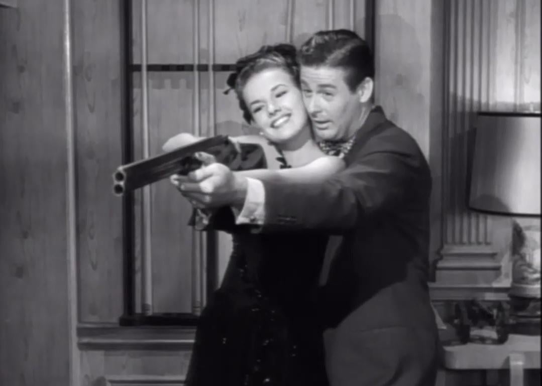 A man showing a woman how to hold a long gun while both are smiling.