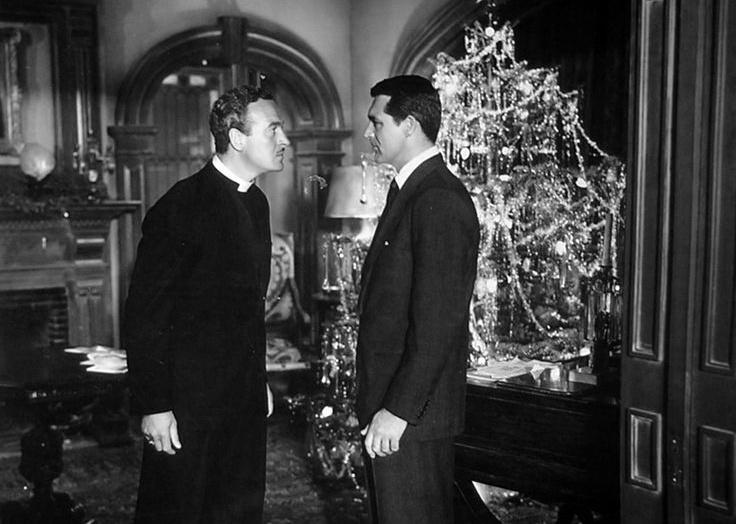 A man and a priest look seriously at one another in front of a Christmas tree.