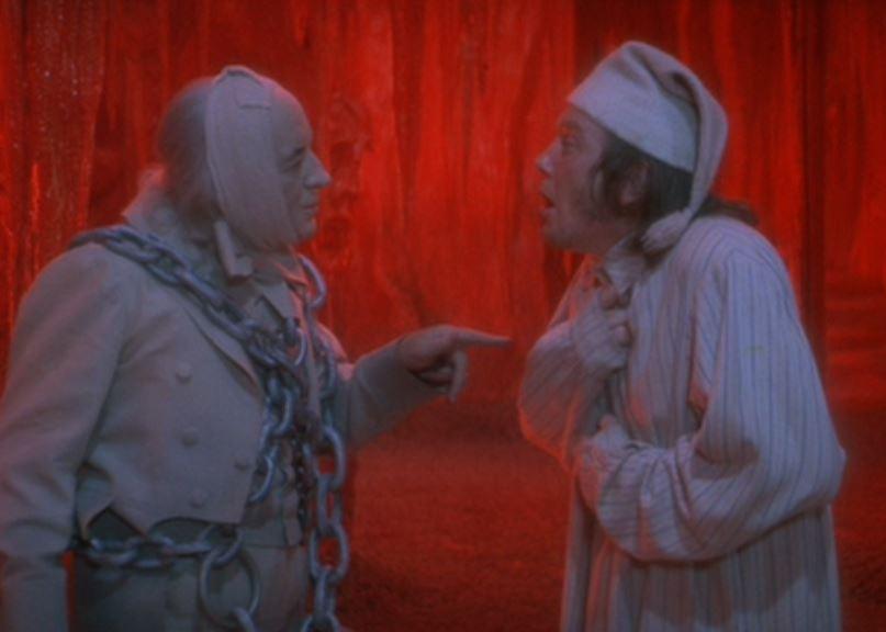 A man in his pajamas talks to a man wrapped in chains in front of a red background.