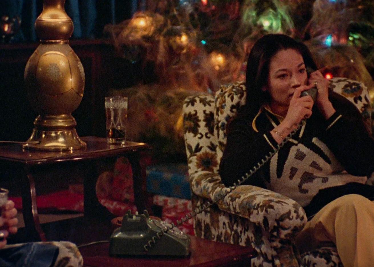A young woman sitting in a floral armchair talking on the phone in front of Christmas lights.
