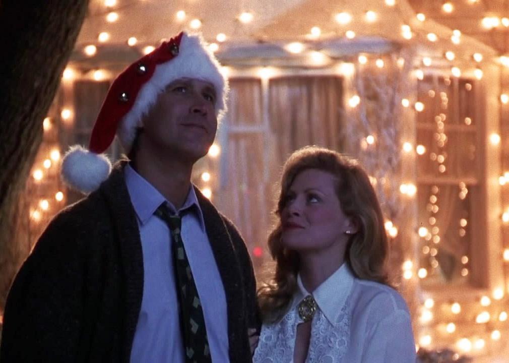 Chevy Chase and Beverly D'Angelo smiling in front of a house of lights.