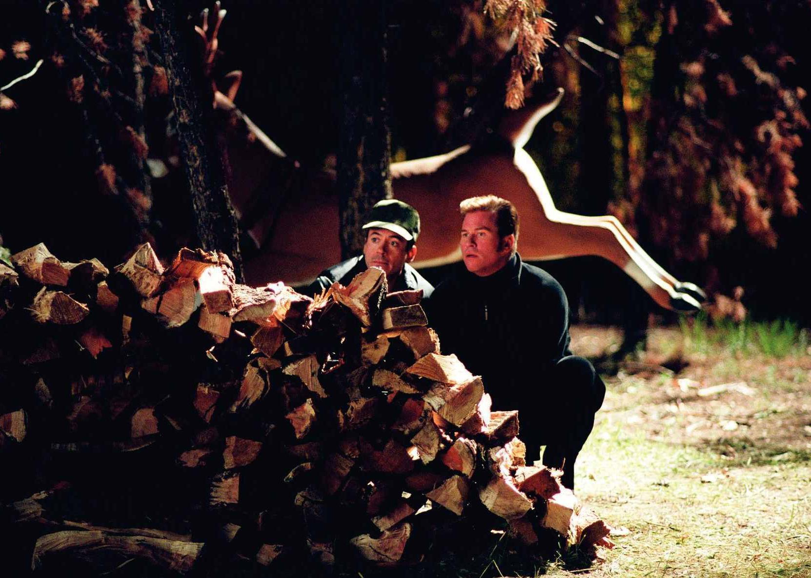 Val Kilmer and Robert Downey Jr. hiding behind a wood pile with a deer jumping in the background.