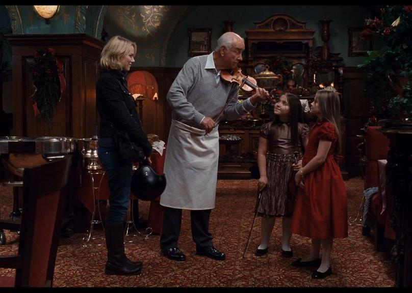 Naomi Watts stands in a restaurant holding a motorcycle helmet while an older man plays a violin for two little girls.