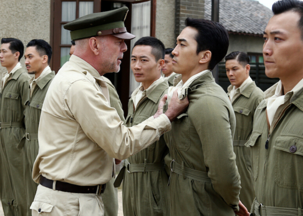 Bruce Willis, dressed in an officer's uniform, grabs another man in uniform by his collar with an angry look on his face while other soldiers look on.