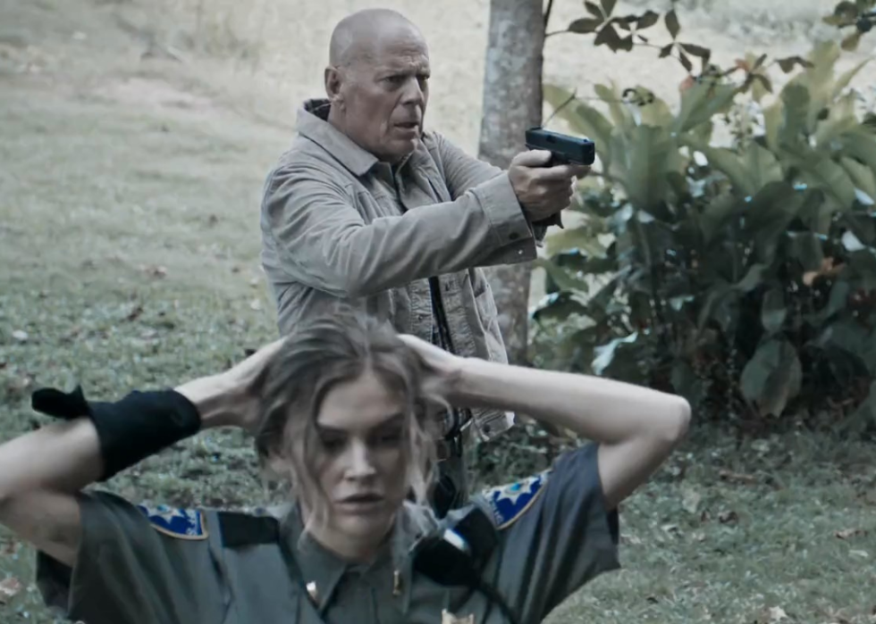 Bruce Willis points a gun at something ahead while a police officer in the foreground has her hands behind her head.