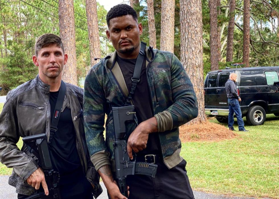 Two men with serious faces patrol with big guns while Bruce Willis is in the background by a black van.