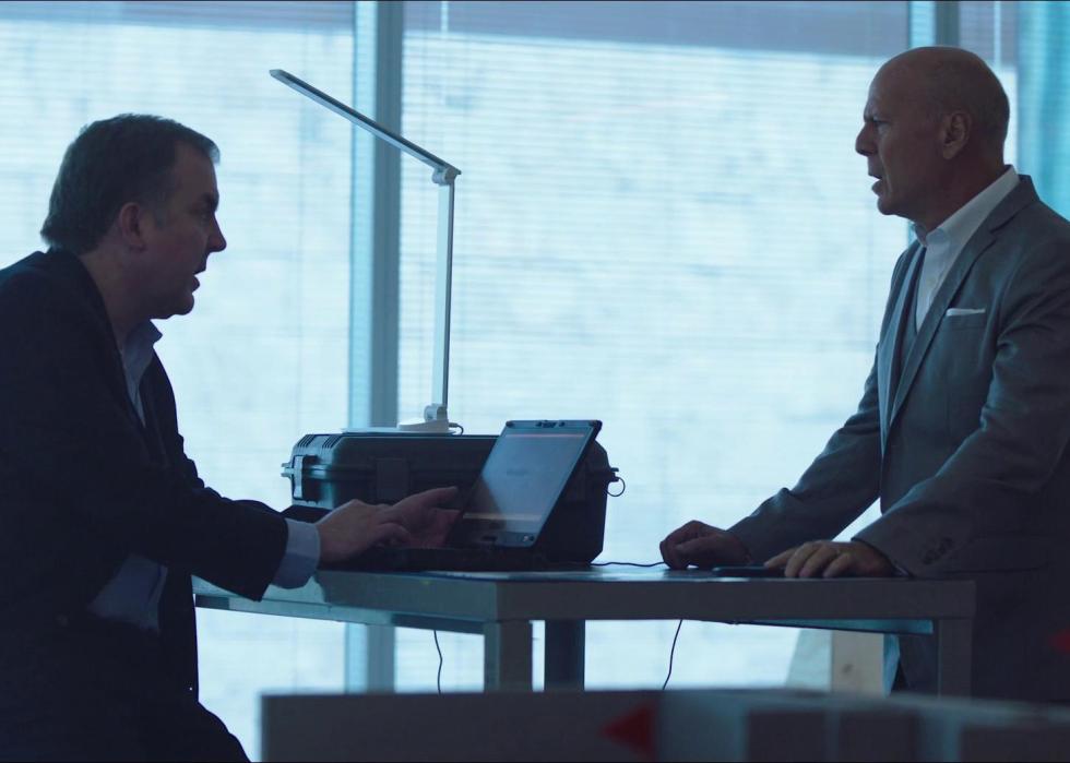2 men talk facing each other over a laptop on a desk next to a silver brief case.