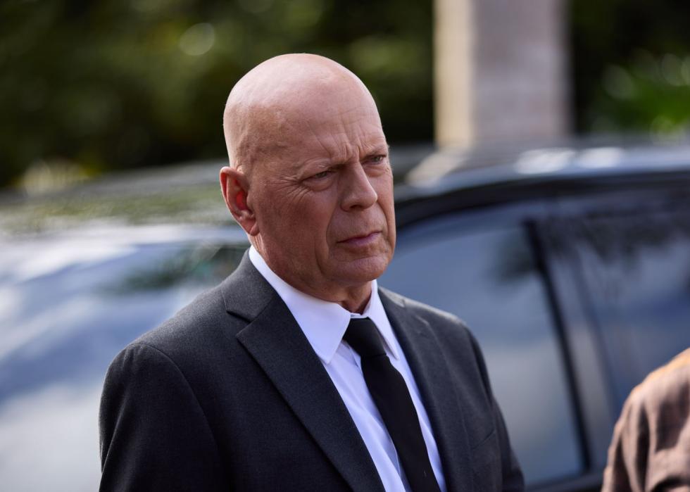 Bruce Willis is dressed in a black suit and tie with a very serious look on his face.