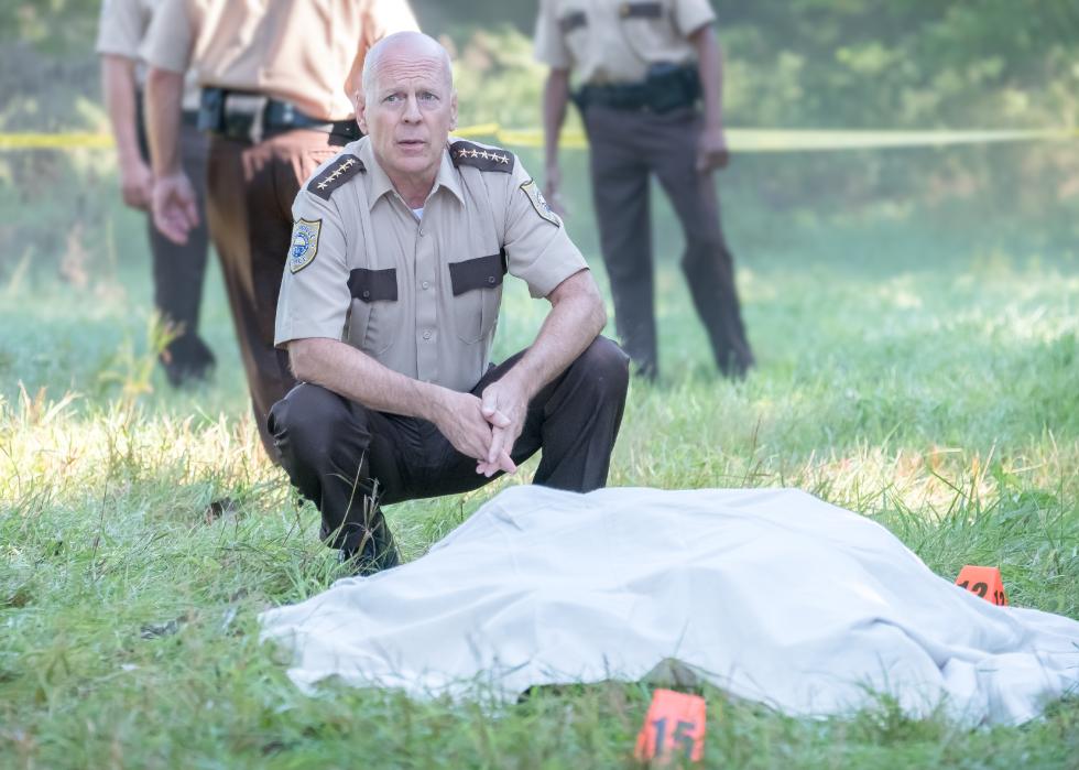 Bruce Willis in a police uniform stands over a covered body in a crime scene.