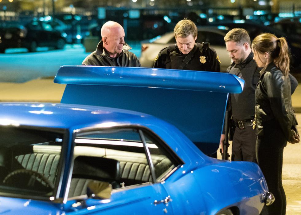 Three men and a woman (all police) look into the trunk of a bright blue vintage car.