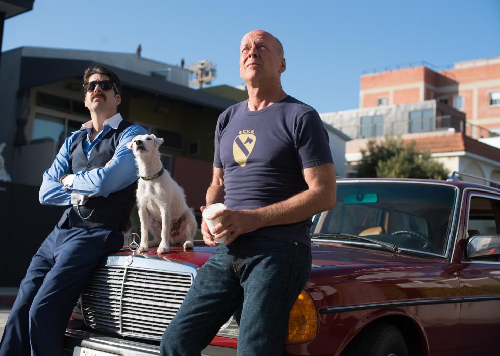 Two men and a little white dog sit on the hood of a classic mercedes while looking up at something.