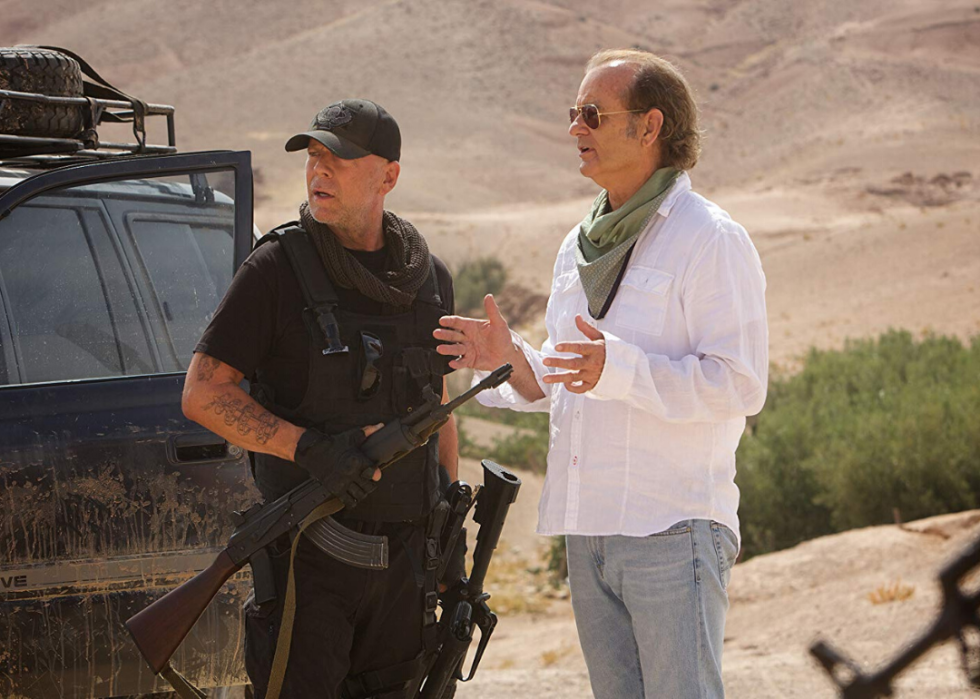 Bruce Willis, dressed in all black with a gun, stands next to Bill Murray, both men looking confusingly at something.