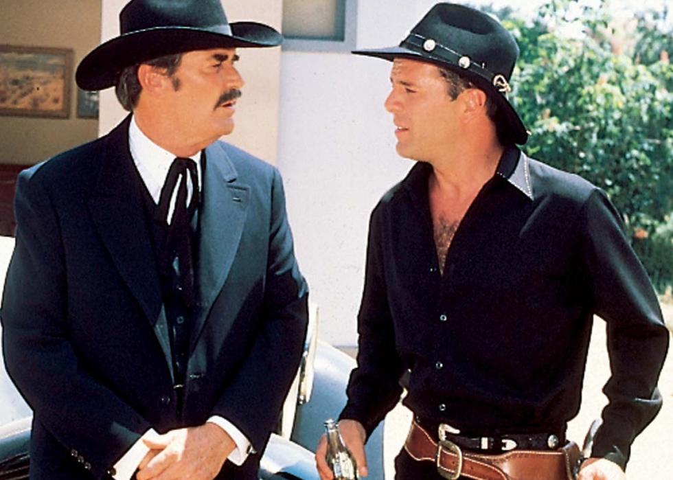 Two men dressed in all black with cowboy hats stand staring at each other.