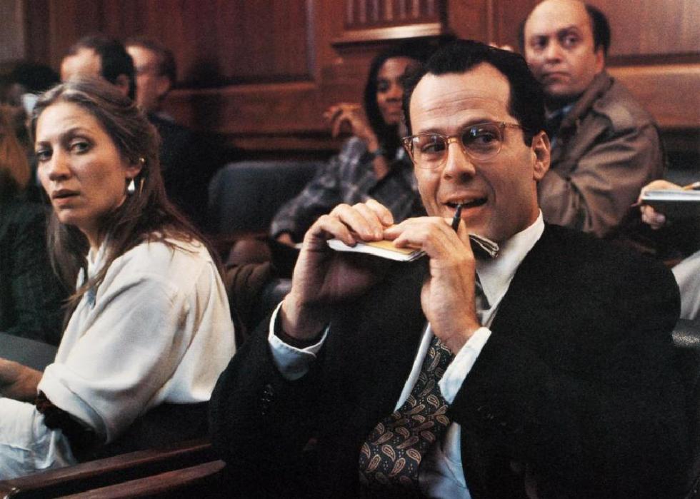 A man dressed in a suit and tie sits in a courtroom smiling while holding a small notepad and pen.