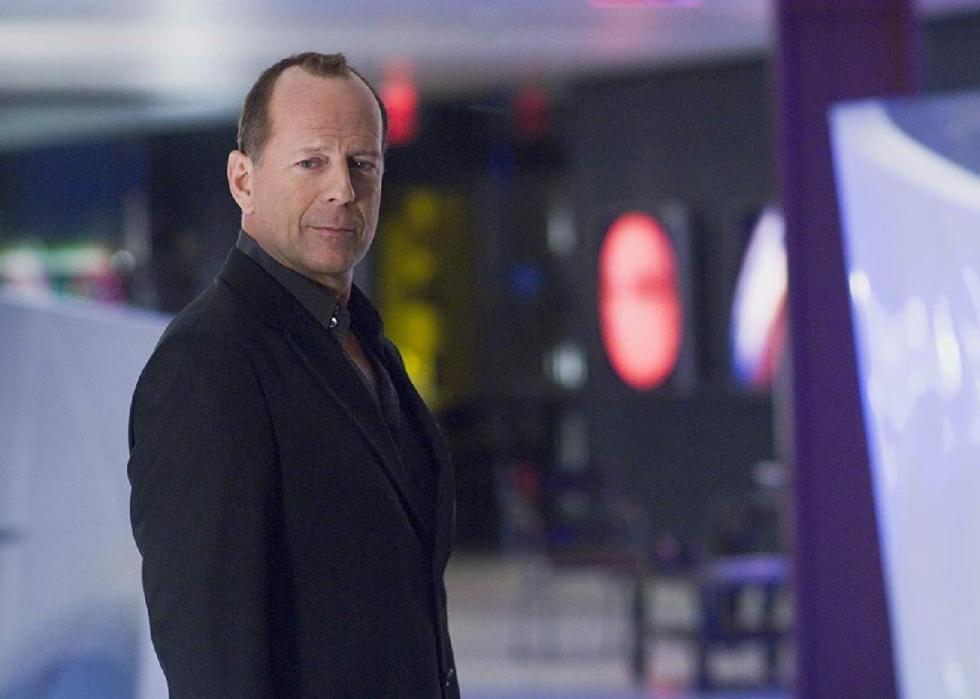 Bruce Willis with a short haircut, smiling and wearing a dark fitted suit.