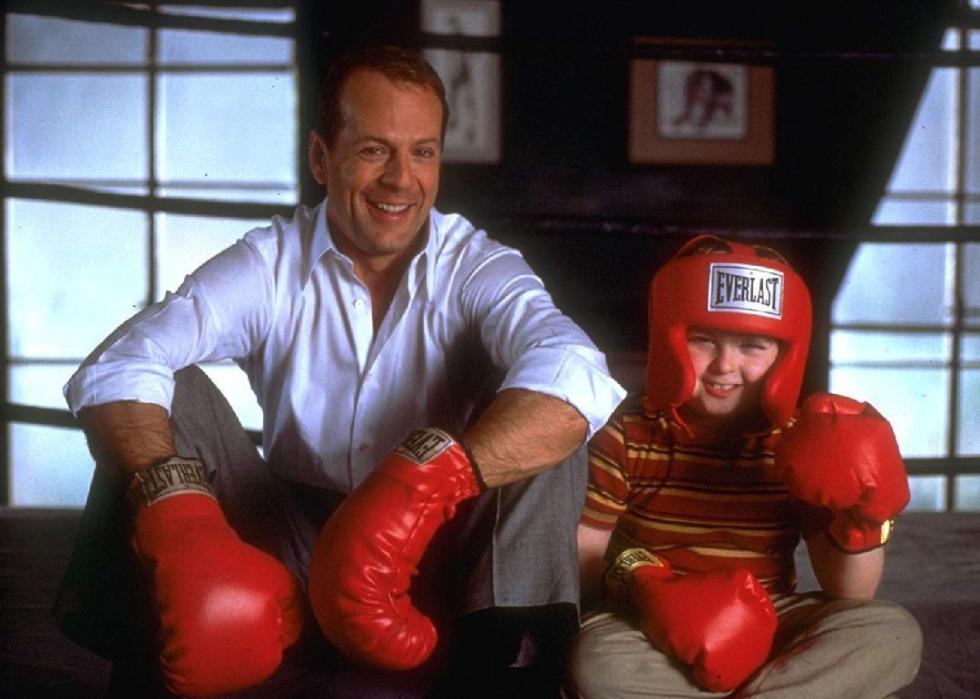 Bruce Willis sits with a little boy, both wearing boxing gloves and smiling.