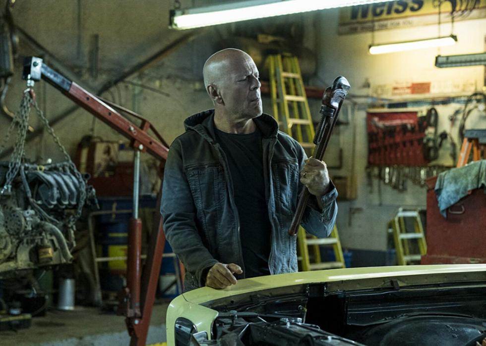 Bruce Willis stands in front of a classic car in a repair shop holding a large wrench.