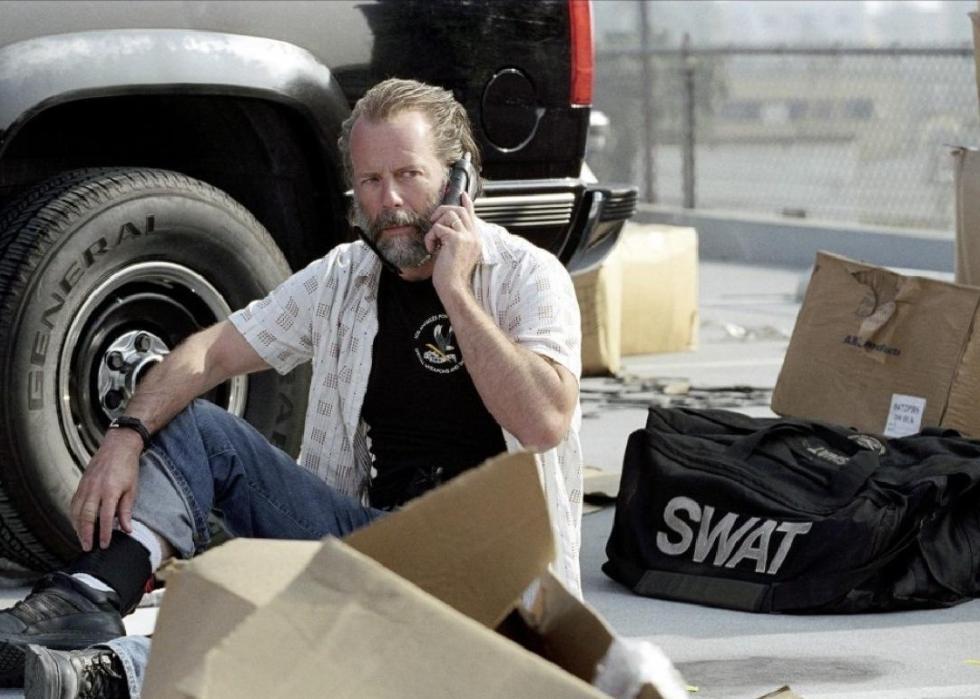 A bearded man on the phone sits next to a black police SUV and a SWAT bag.