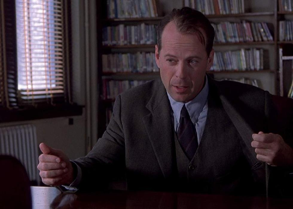 Bruce Willis sits at a desk in front of a bookcase gesturing with his hands.