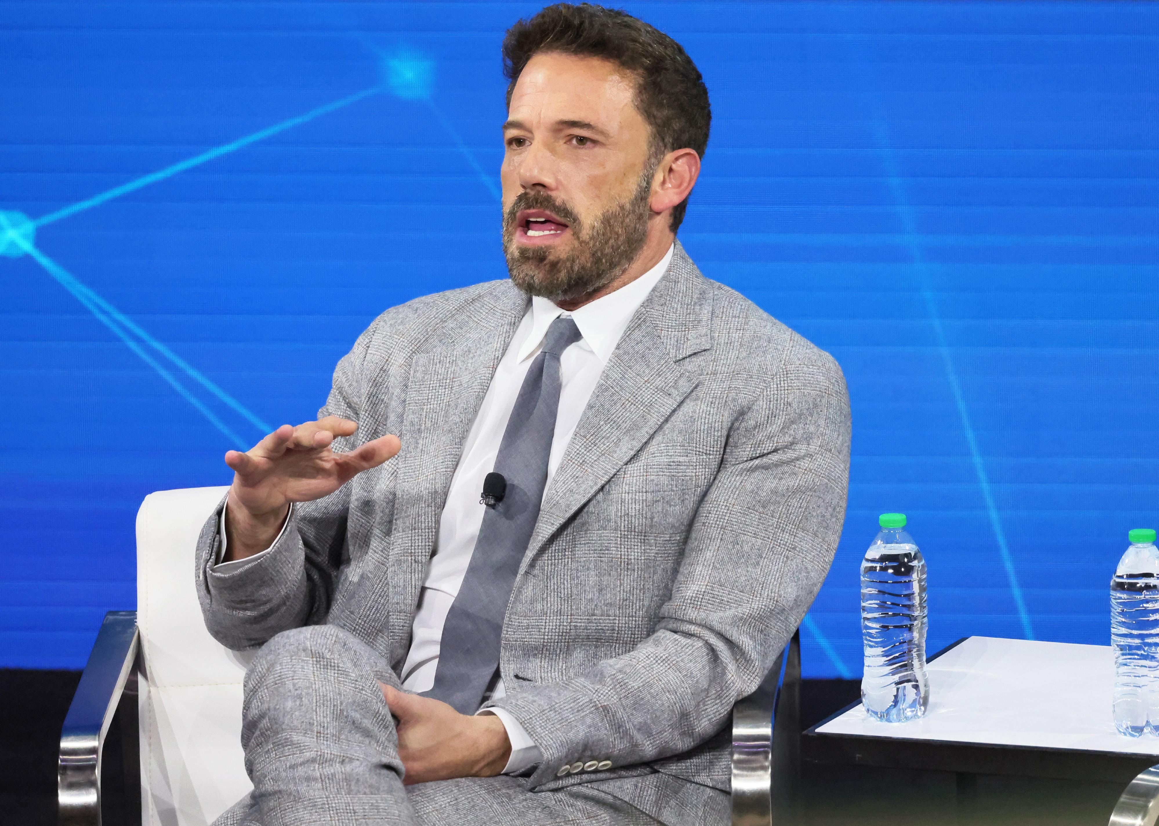 Ben Affleck in a grey suit onstage in front of a blue background.