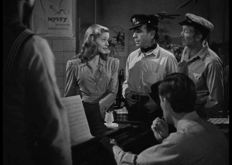 Black and white image of two men with fishing hats on talk with a blonde woman in next to a piano.