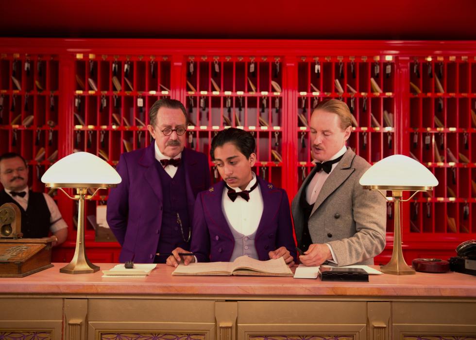 Hotel employees dressed in colorful suits look over a guest book at reception.