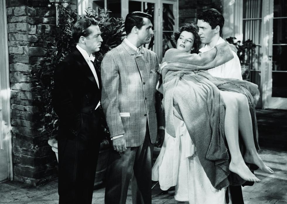 One man carries a woman in a robe while he talks to two other men.