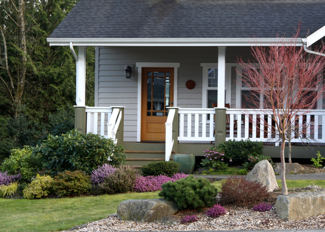 A small gray house with a front porch and blooming plants in front.