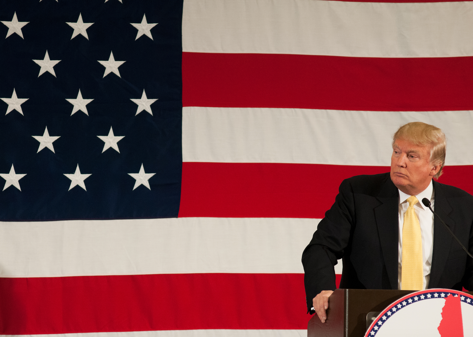 Donald Trump in a black suit at a podium in front of an American flag.