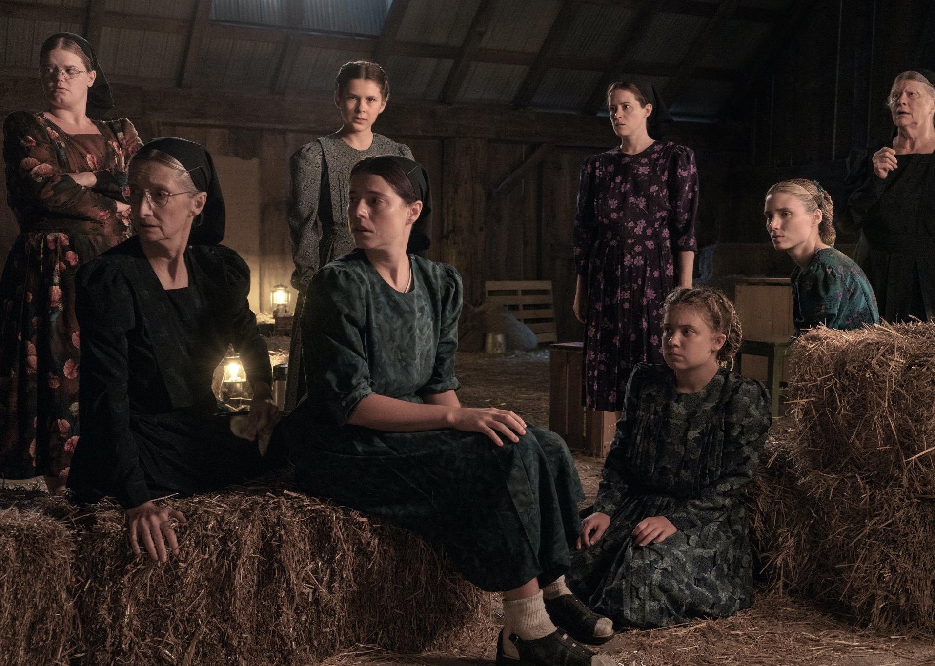 A group of women with worried looks on their faces sit on hay bales in a barn.