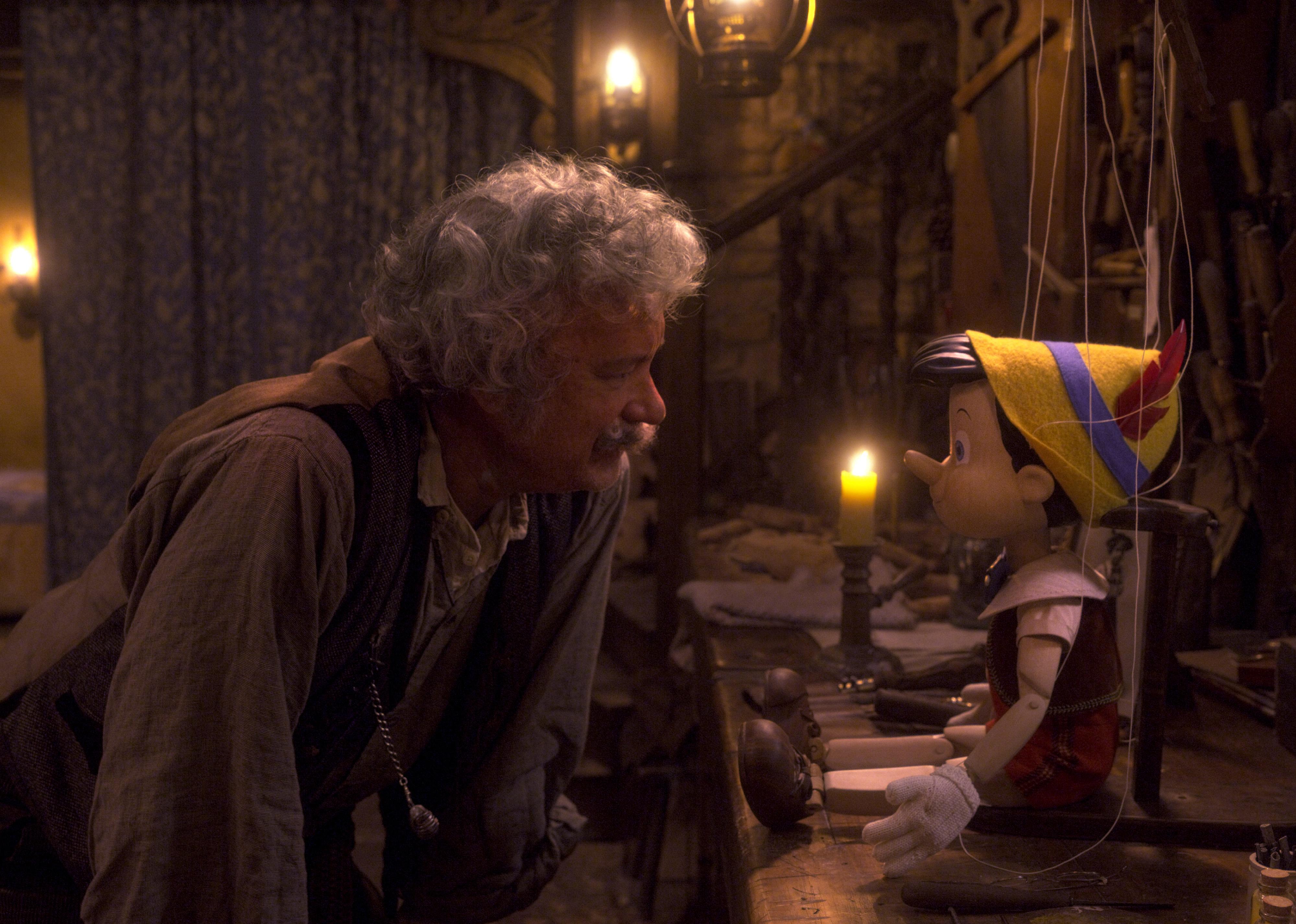 An older man (Tom Hanks as Geppetto) leaning over a table to look at a puppet of a little boy (Pinocchio).