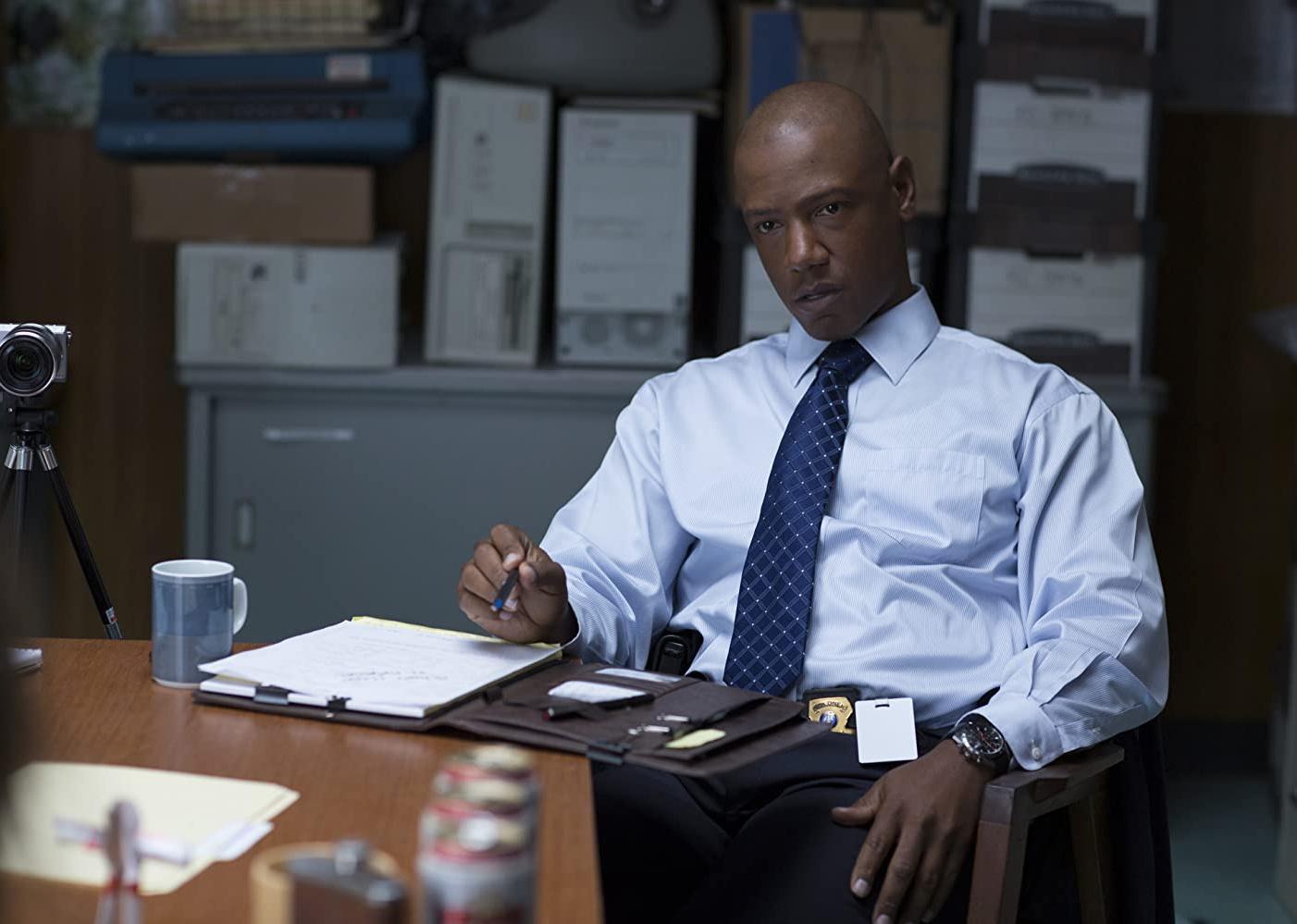 A police detective wearing a blue button down shirt and tie at a desk.