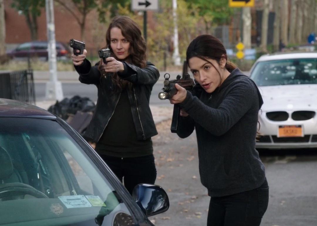 Two women wearing all black in the street point guns at someone.