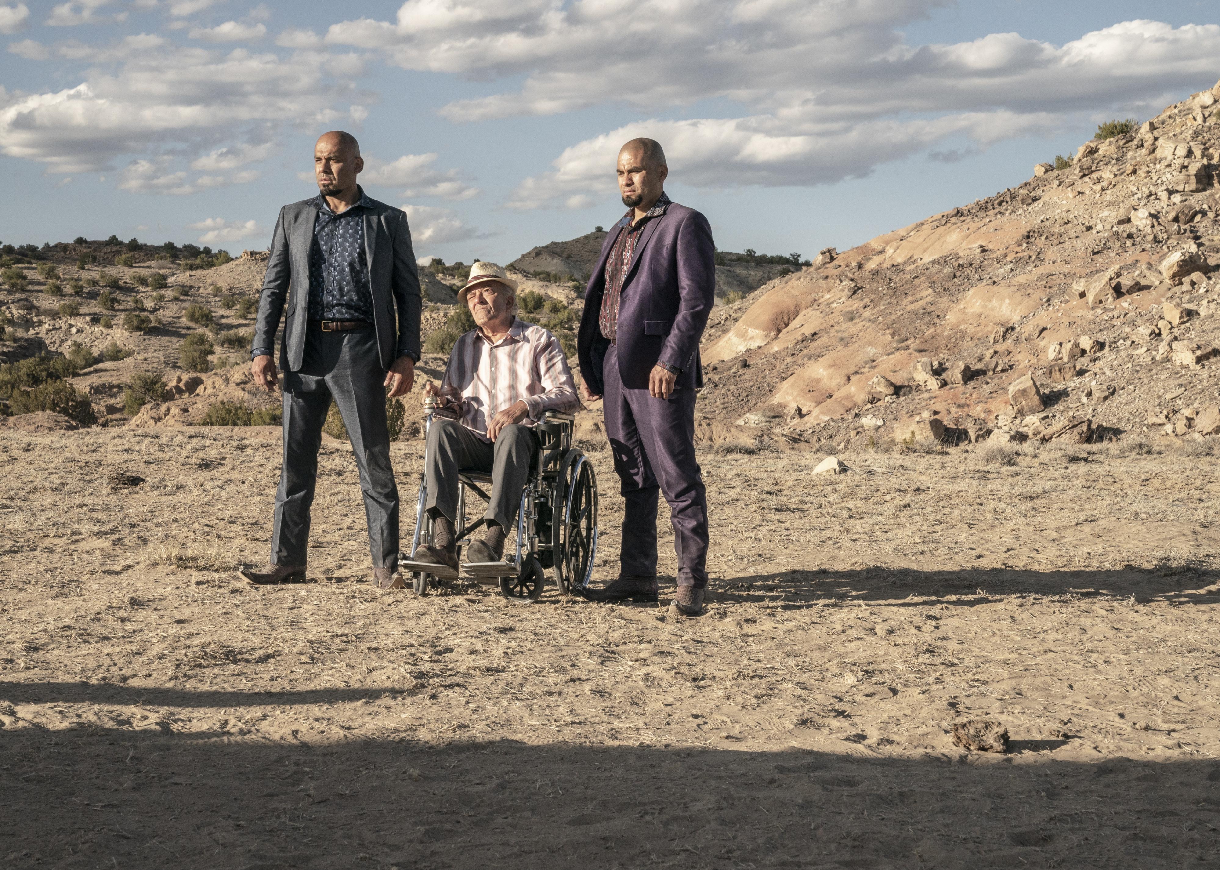Two identical men in suits and cowboy boots stand with a man in a wheelchair in between them in the desert.