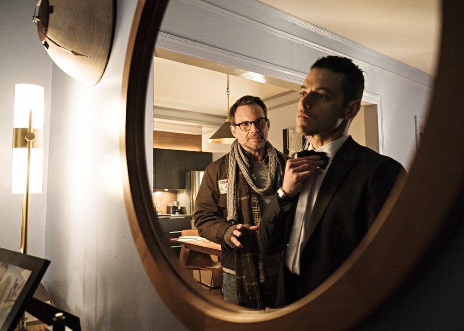 Rami Malek looks in the mirror, wearing a tuxedo, with Christian Slater next to him.