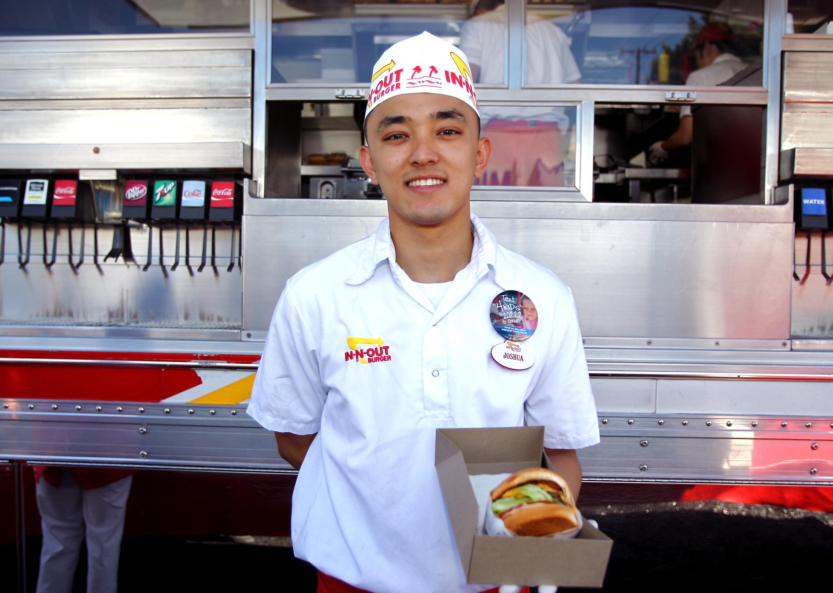 An In-N-Out Burger employee in a white shirt and hat holding a burger in a box.