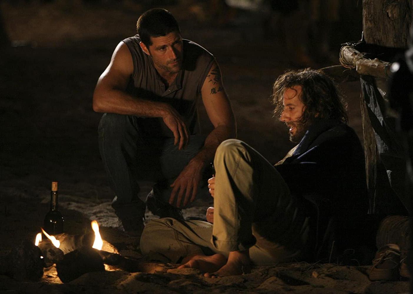 Matthew Fox and another man sit by a campfire at night.