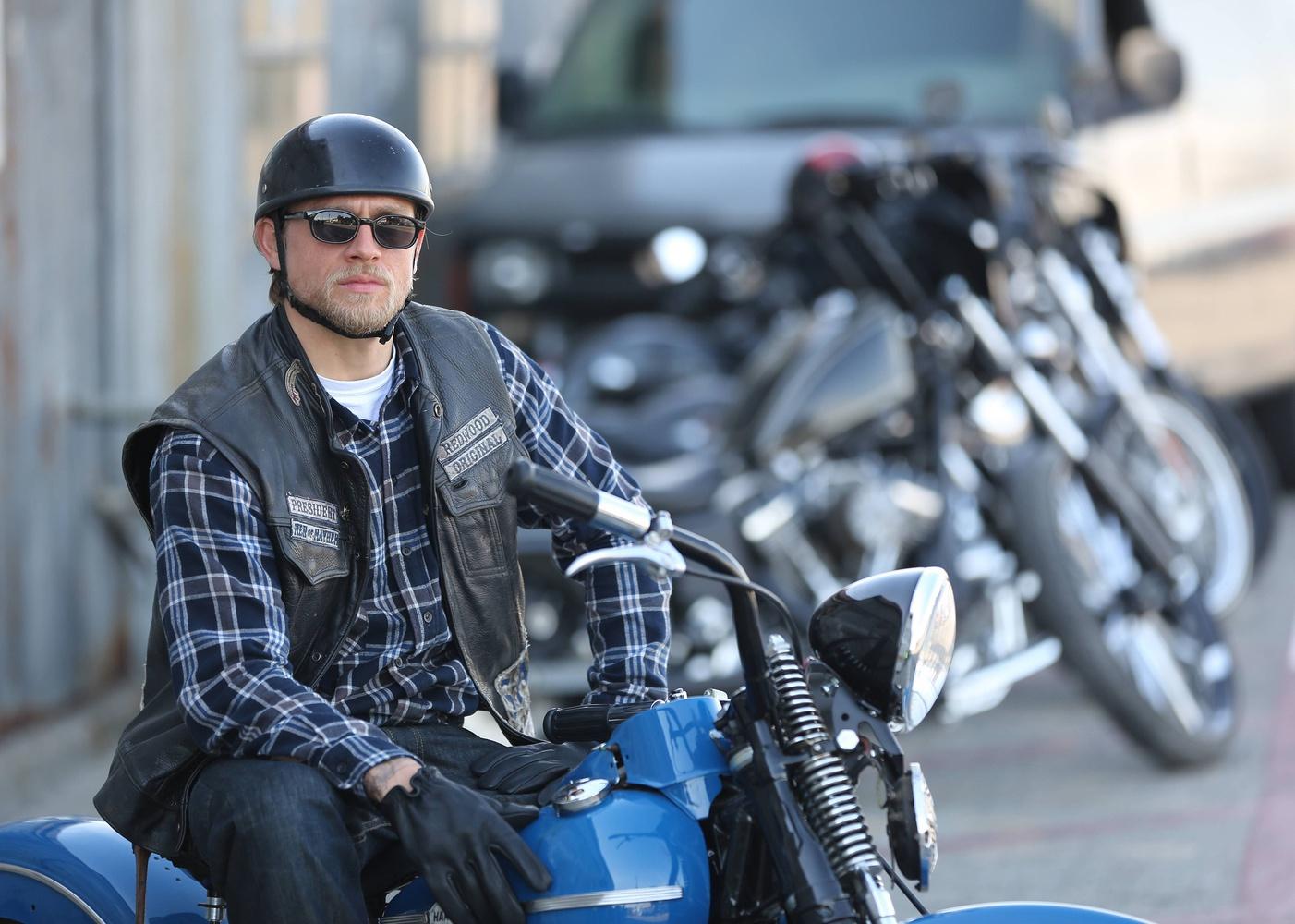 Charlie Hunnam sitting on a motorcycle in a plaid shirt and leather vest.