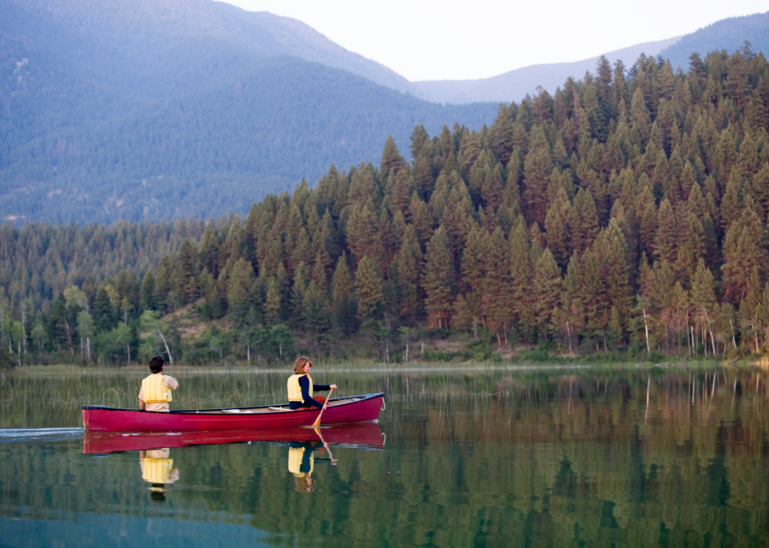 Two people row in a red canoe on a calm river surrounded by forest and mountains. 