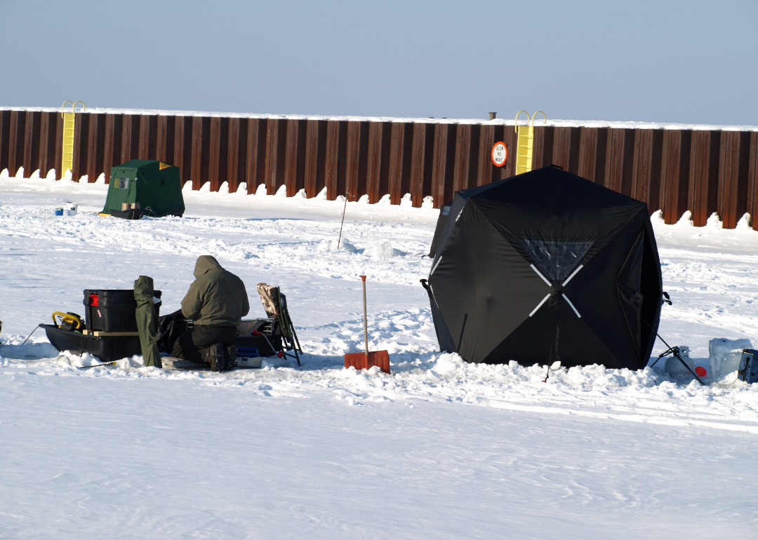 A person bundled up, surrounded by ice fishing supplies. 