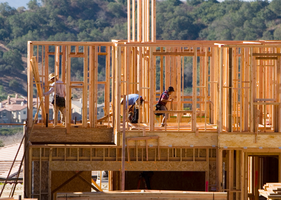A three-person construction team works on the frame of a two-story house in the hills.